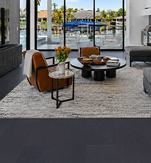 Luxury Black Real Leather Flooring In A Waterfront Living Room With Gray Sectional Sofa, Textured Area Rug And Large Glass Doors Overlooking The Water.